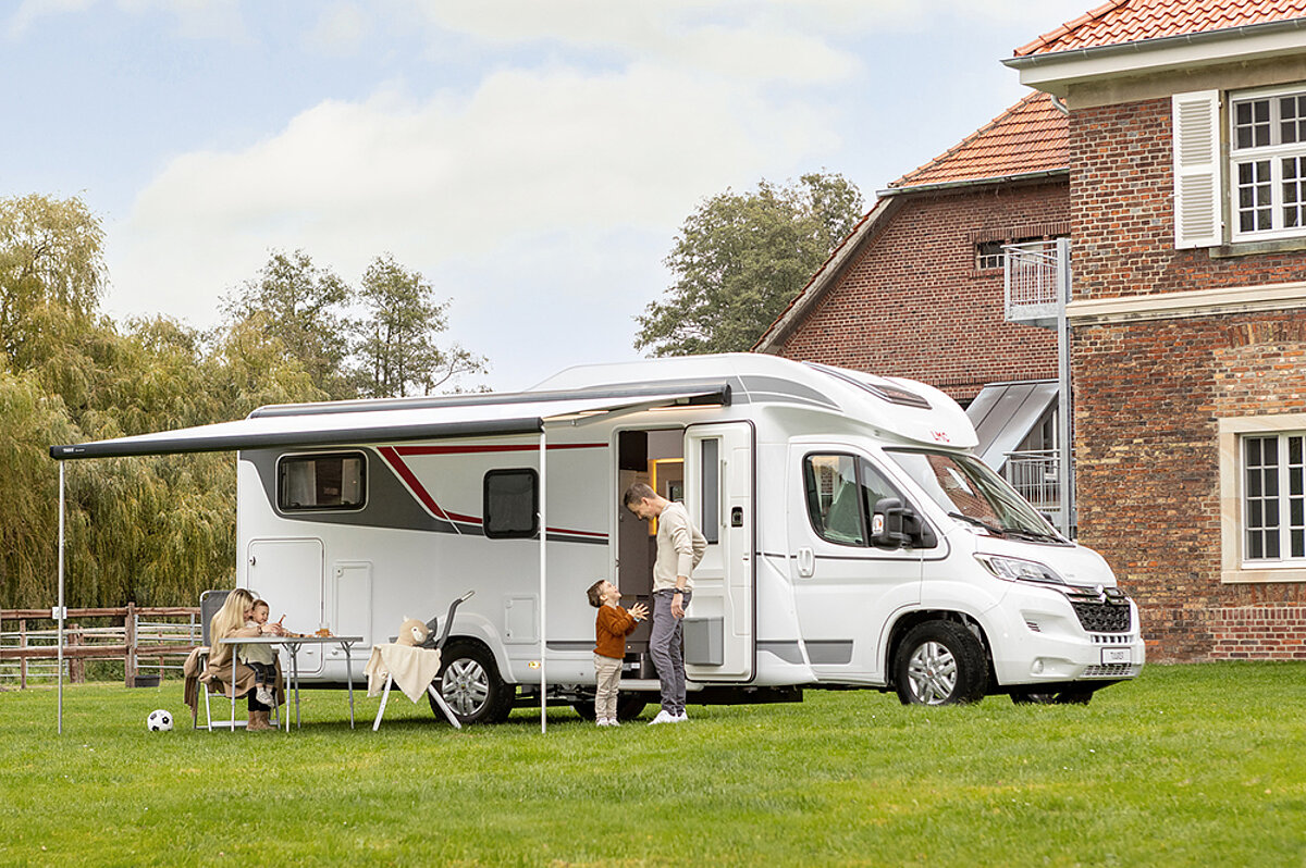 Ein weißes Wohnmobil mit aufgestelltem Vordach auf einer Wiese vor einem Backsteinhaus. Davor stehen und sitzen eine Familie mit zwei kleinen Kindern.
