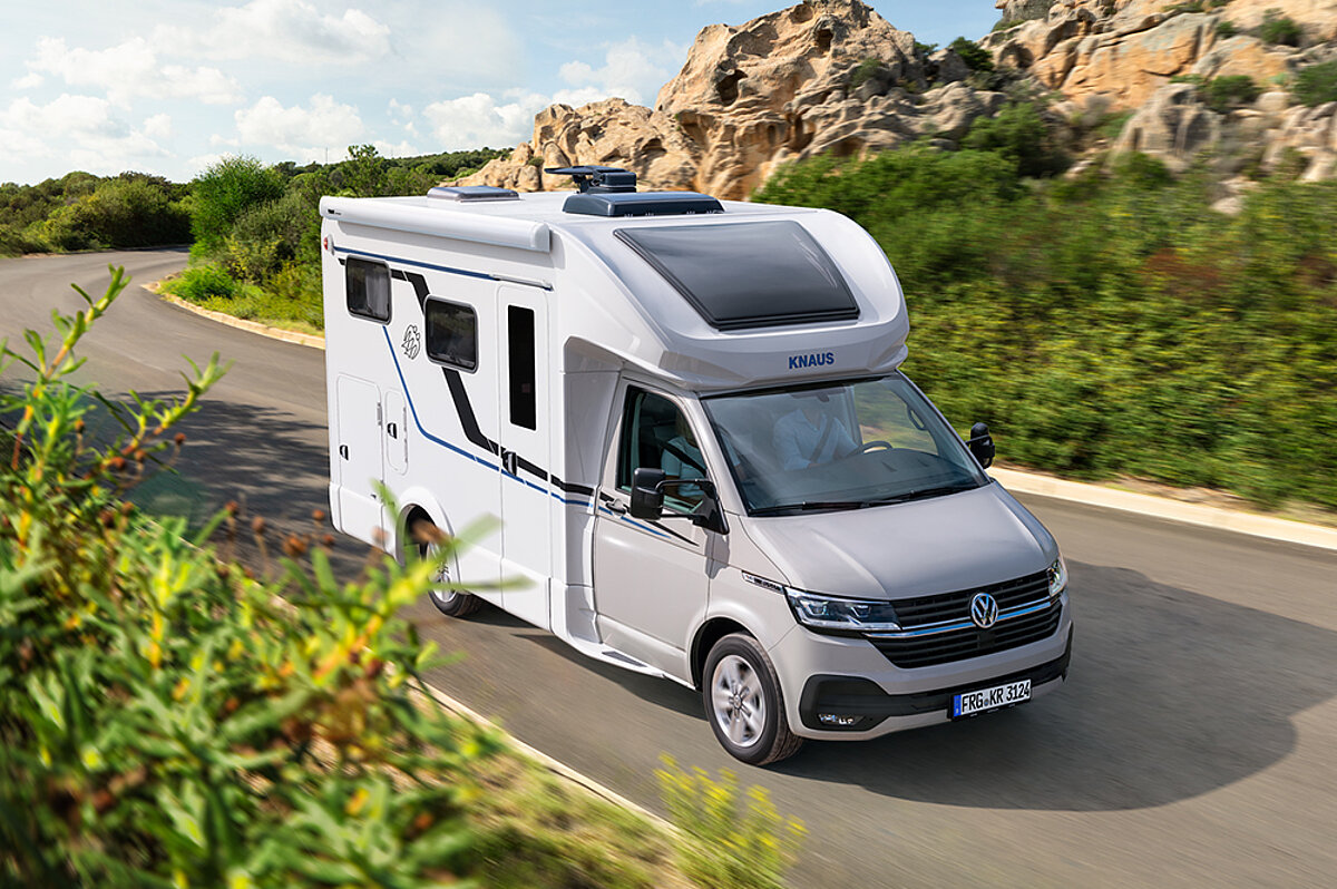 Wohnmobilurlaub verbindet Abenteuerlust mit dem Komfort eines mobilen Zuhauses. Wohnmobil auf einer Straße in einer Landschaft mit grünen Büschen im Vordergrund.