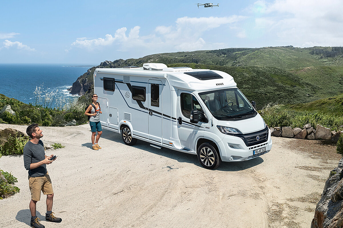 Urlaub mit dem Wohnmobil bietet den Luxus, überall ein Zuhause zu haben, egal wohin die Reise geht. Ein Wohnmobil steht auf einem Parkplatz mit Blick aufs Meer. Zwei Personen sind im Vordergrund zu sehen.