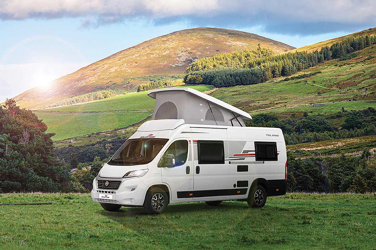 Im Van die Freiheit spüren, den Alltag hinter sich zu lassen und die Welt zu entdecken. Ein weißer Campervan mit aufgestelltem Dach auf einer grünen Wiese und einer bergigen Landschaft im Hintergrund.