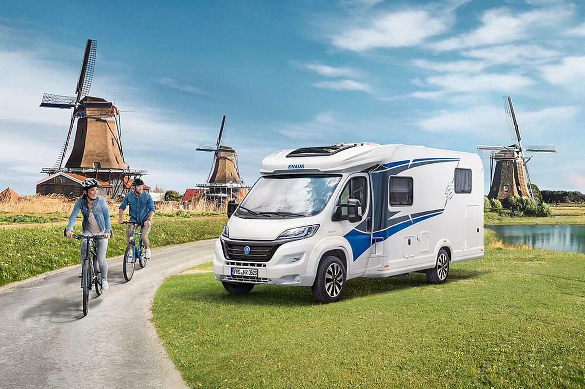 Beim Reisen mit dem Wohnmobil wird jeder Tag zu einem neuen Erlebnis in der freien Natur. Weißes Wohnmobil mit blauen Details steht auf einer Wiese vor drei Windmühlen. Zwei Personen auf Fahrrädern fahren an dem Wohnmobil auf einer Straße vorbei.