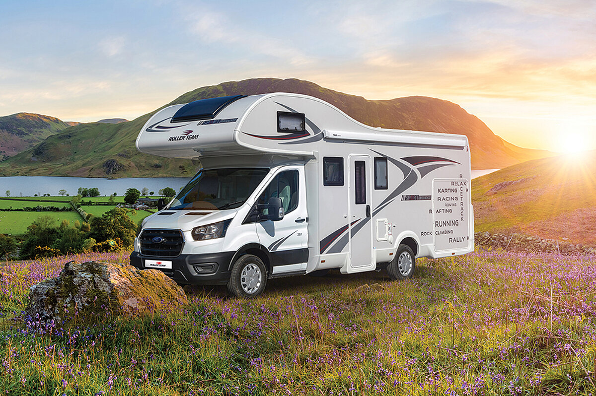 Wir genießen die Meeresbrise und das Salz in der Luft, während unser Wohnmobil uns bequem überallhin begleitet. Ein Wohnmobil mit Alkoven steht auf einer Blumenwiese. Im Hintergrund ist eine Landschaft mit Bergen und Meer zu sehen.