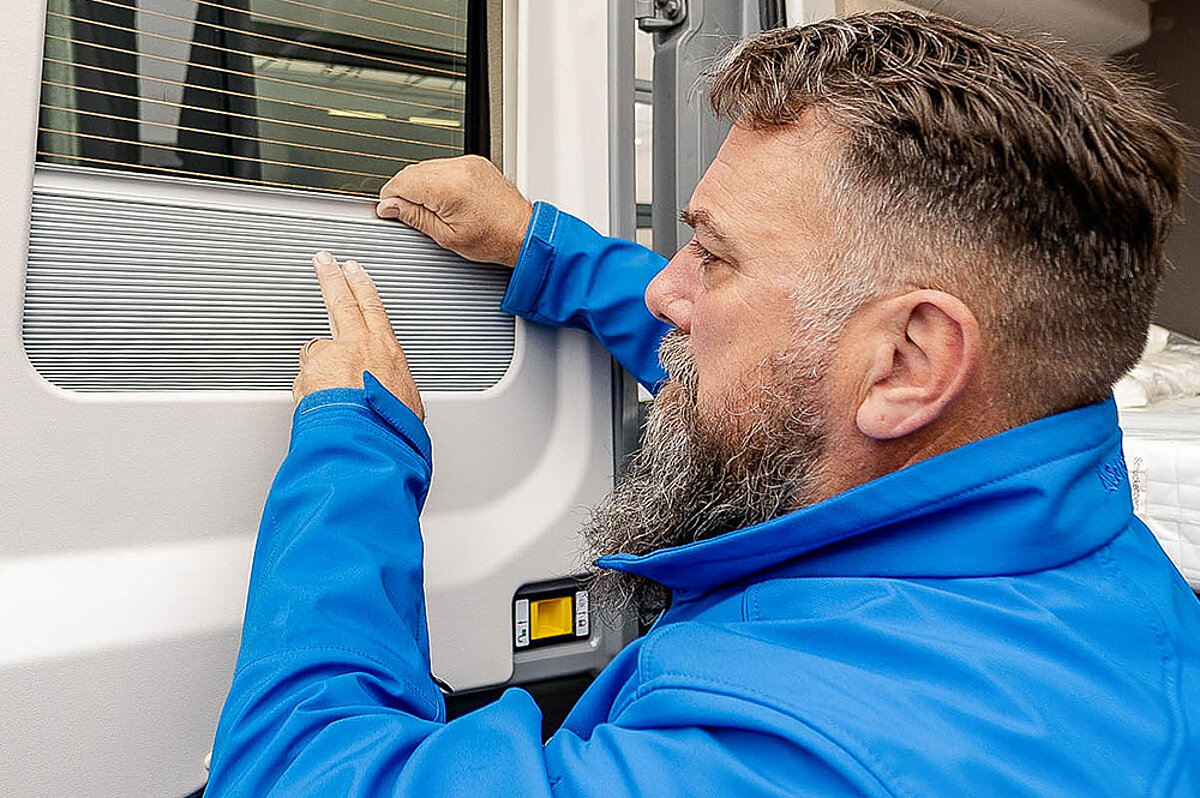 Unsere Experten stehen Ihnen mit Rat und Tat zur Seite, um Ihren Campingurlaub perfekt zu gestalten. Mann in blauer Jacke korrigiert eine Jalousie an einem Fenster.
