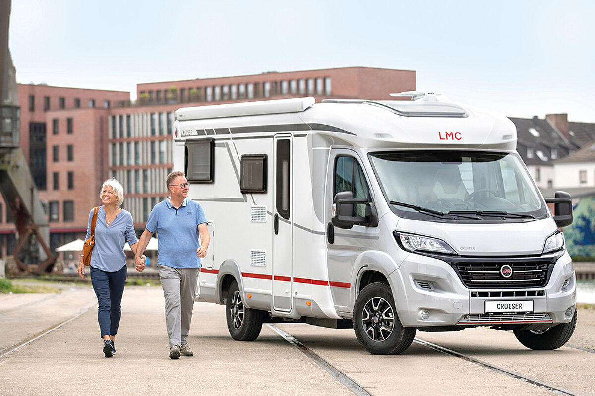 Ein weißes Wohnmobil steht in einer Stadt. Ein Pärchen aus einem Mann und einer Frau gehen händchenhaltend an dem Wohnmobil vorbei.