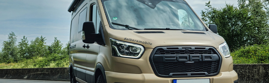 Frontansicht eines goldfarbenen Campervans mit Natur im Hintergrund.
