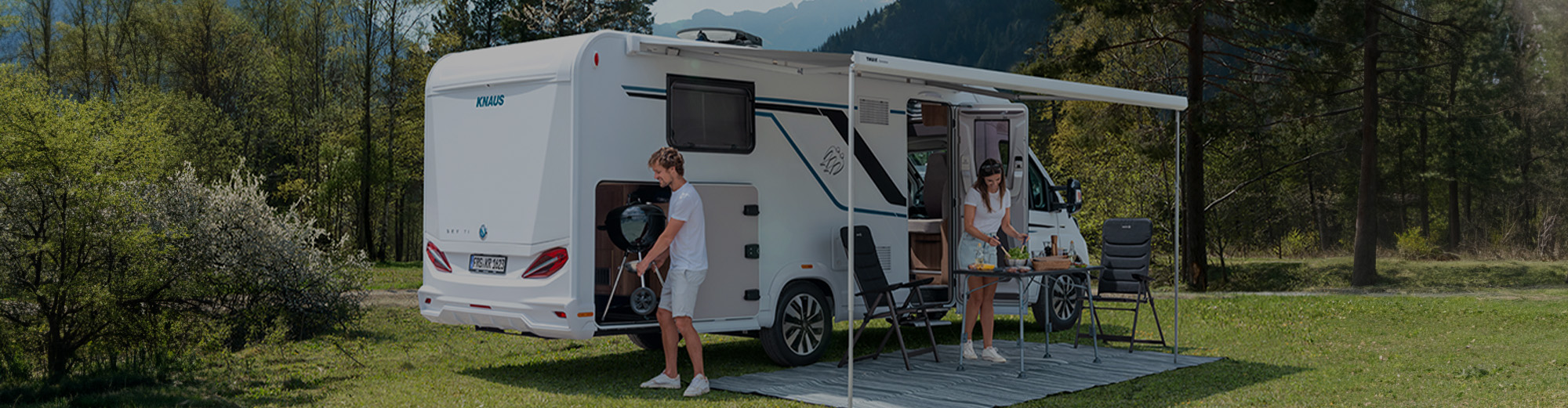 Ein Wohnmobil in einer Landschaft mit geöffneter Tür und aufgestelltem Vordach. Zwei Personen befinden sich vor dem Wohnmobil.