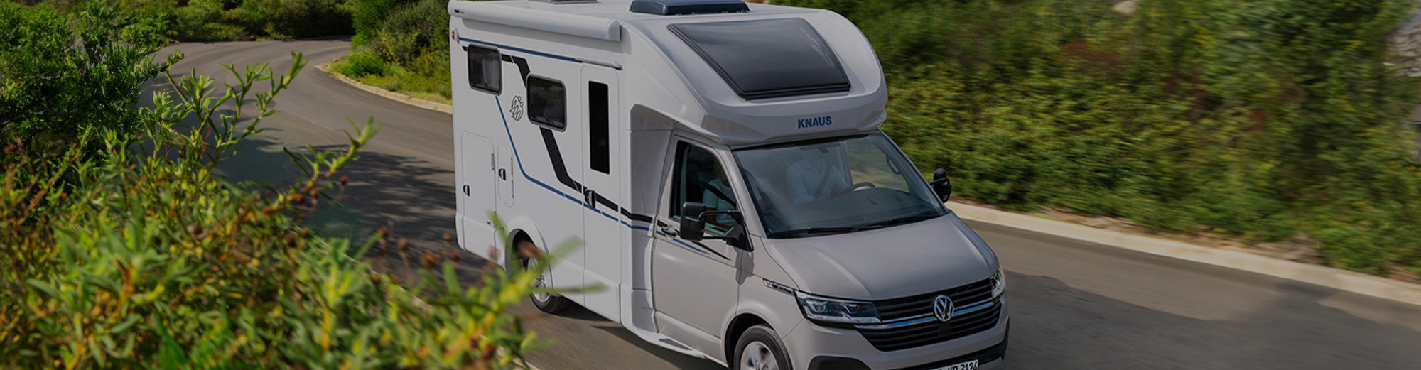Ein Wohnmobil auf einer Landstraße mit vielen Büschen im Vordergrund.
